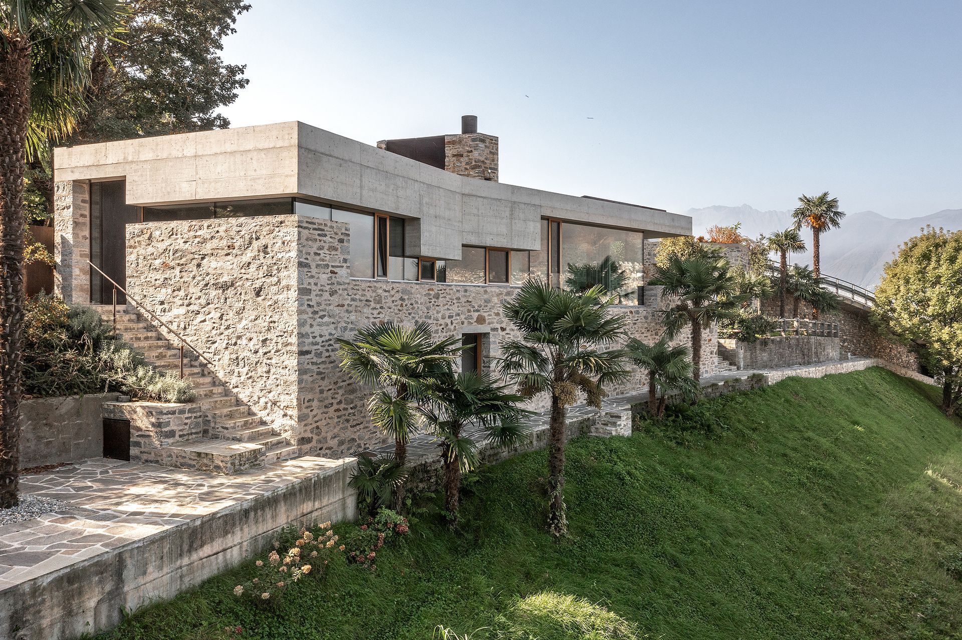 Moderna casa in pietra su una collina con tetto in cemento e palme.