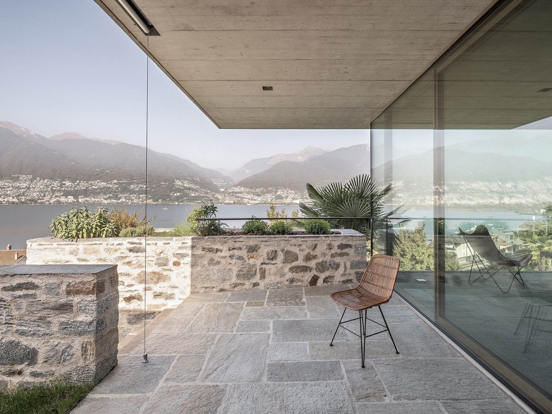 Patio in pietra con sedia e vista sul lago e sulle montagne attraverso una parete di vetro.