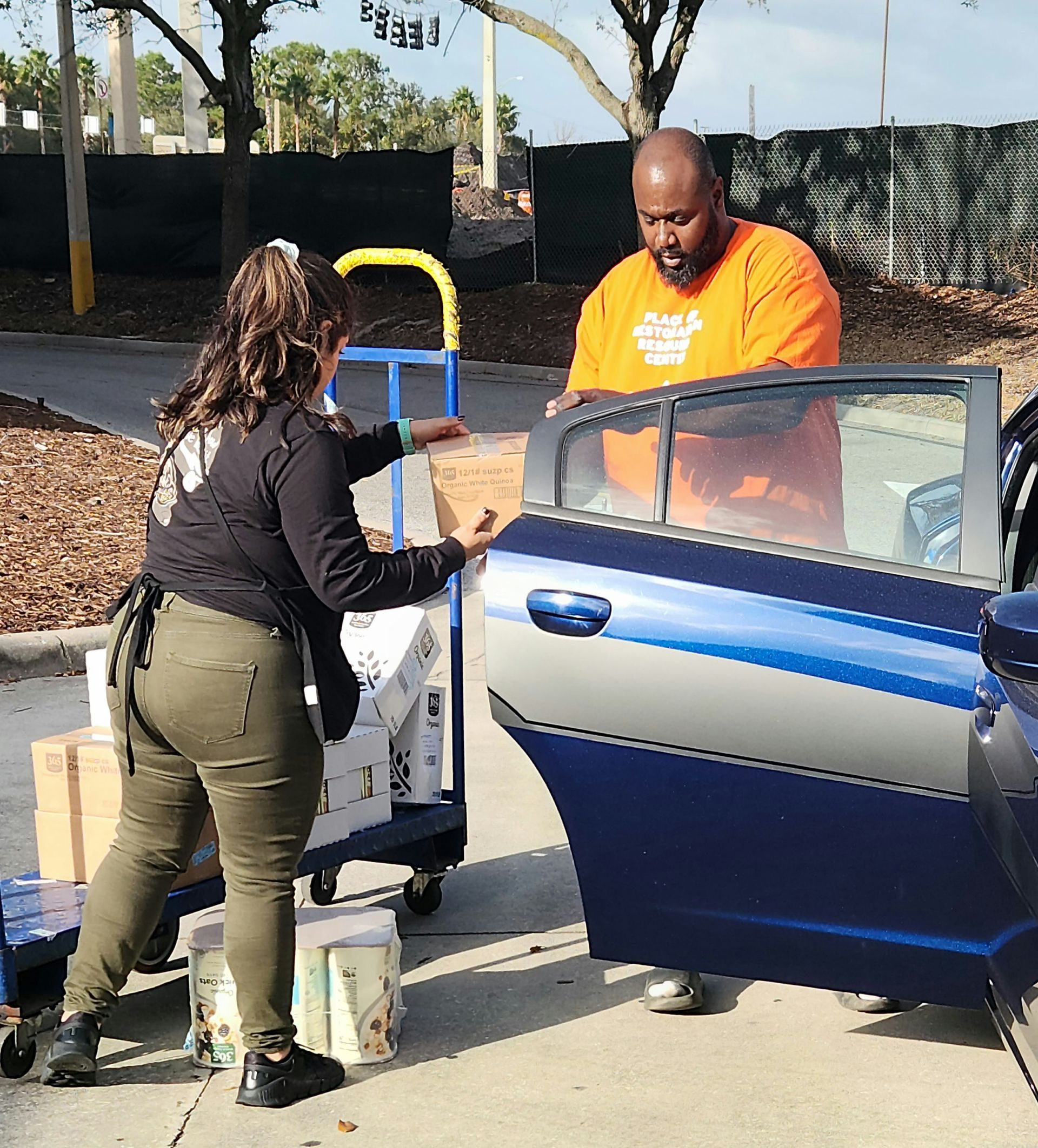 Woman loads items from a cart into a blue car trunk as a man in orange shirt assists outside.