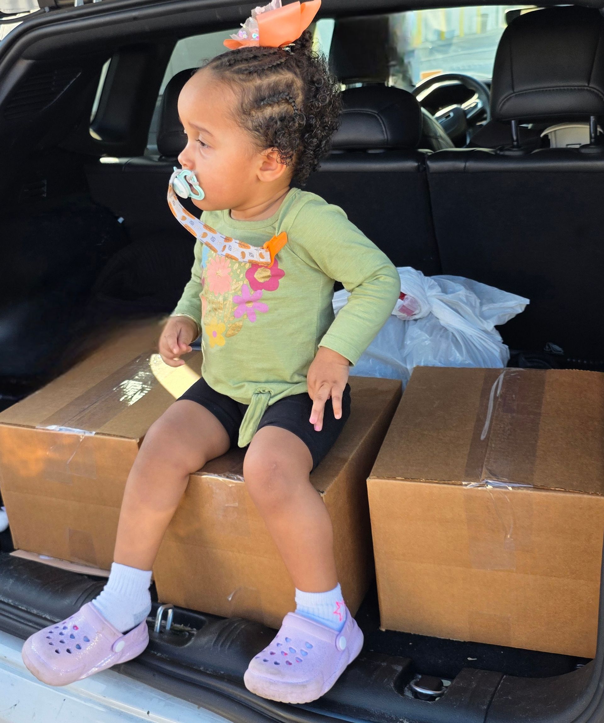 Toddler sitting on cardboard boxes in the back of an SUV, wearing green top and purple shoes.