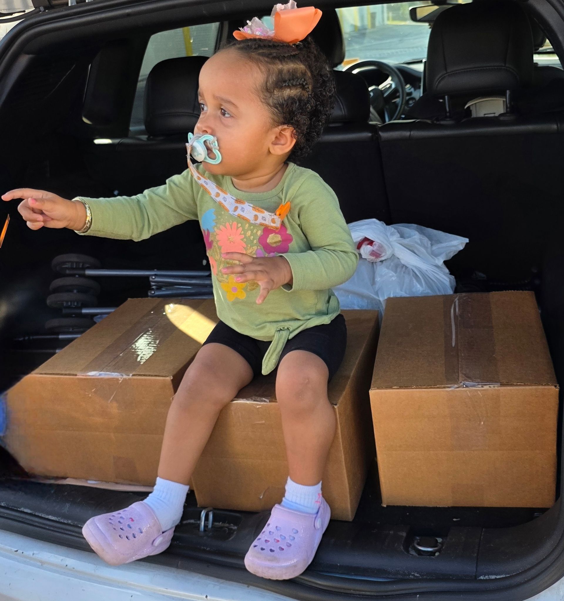 Toddler sitting in an SUV trunk on cardboard boxes, wearing a pacifier and reaching forward.