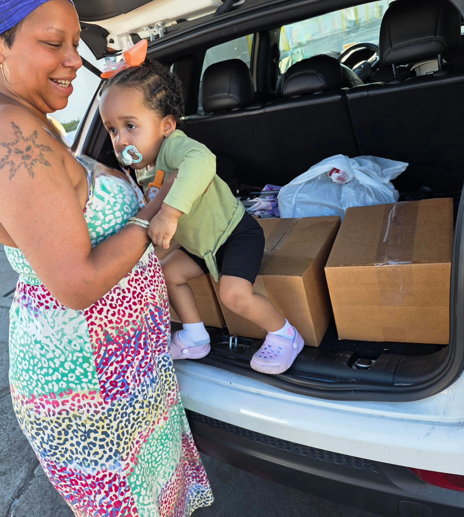 Woman holding a baby beside cardboard boxes in the open trunk of an SUV