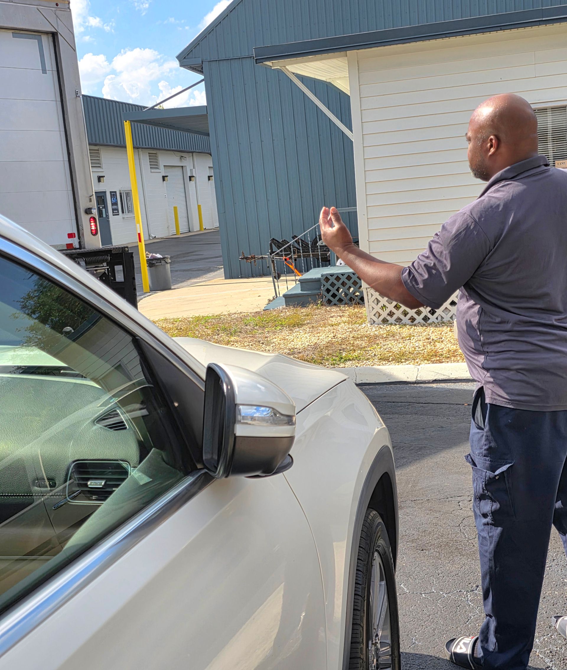 Man standing beside a white car in a parking lot, gesturing toward a building