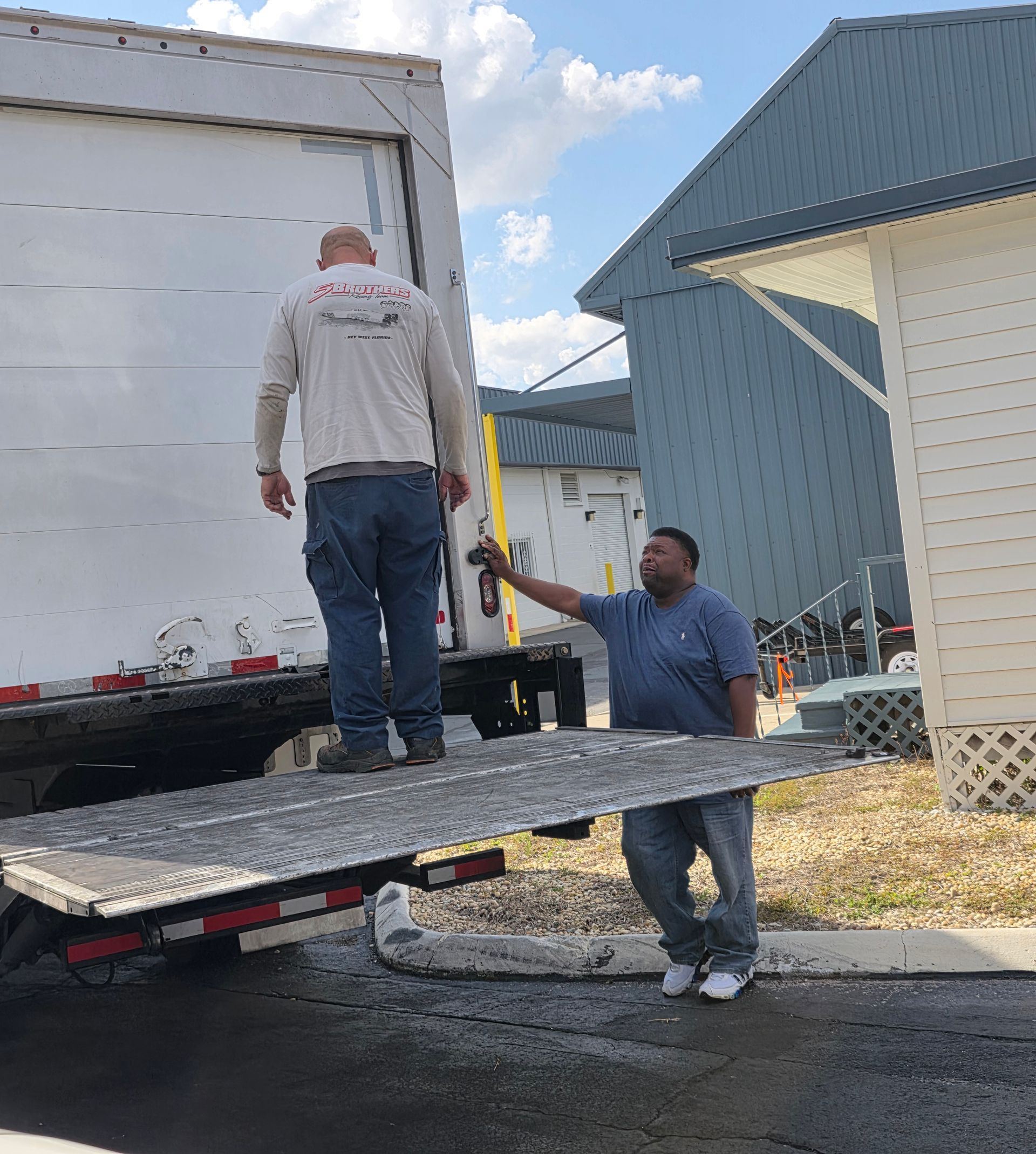 Two men load a white box truck from a ramp beside a house under a blue sky.