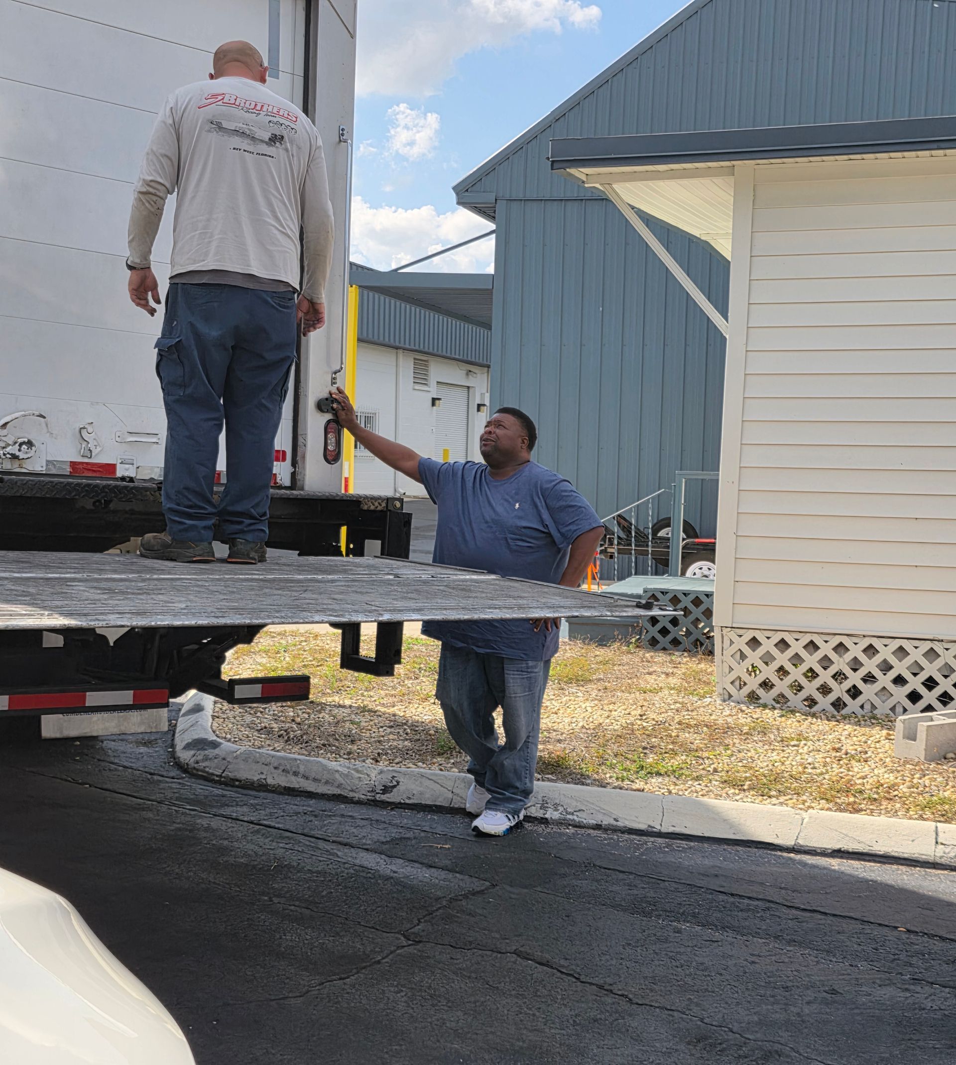 Two men unloading equipment from a truck beside a blue building on a sunny day