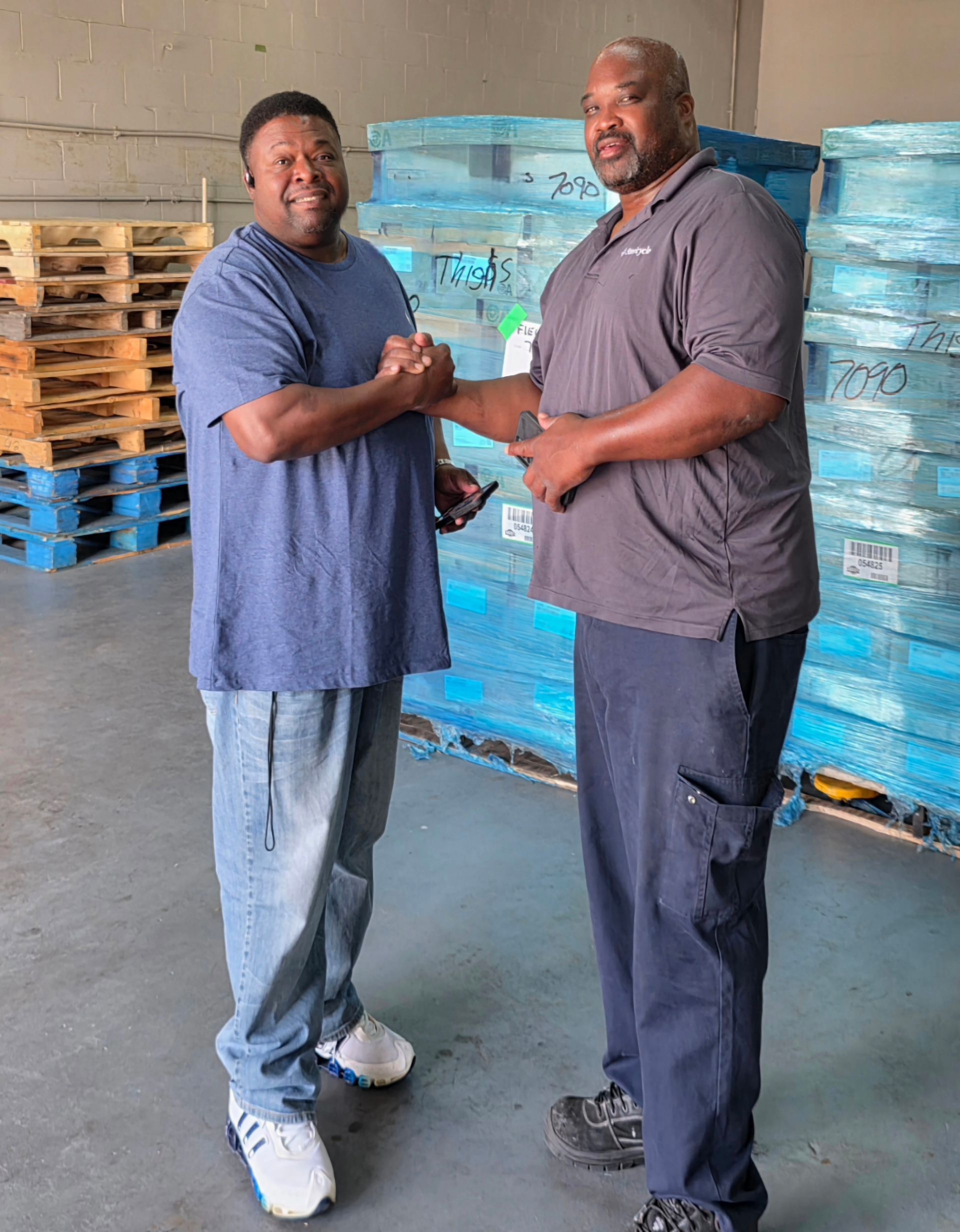 Two men shaking hands in a warehouse with stacked pallets and blue-wrapped goods.