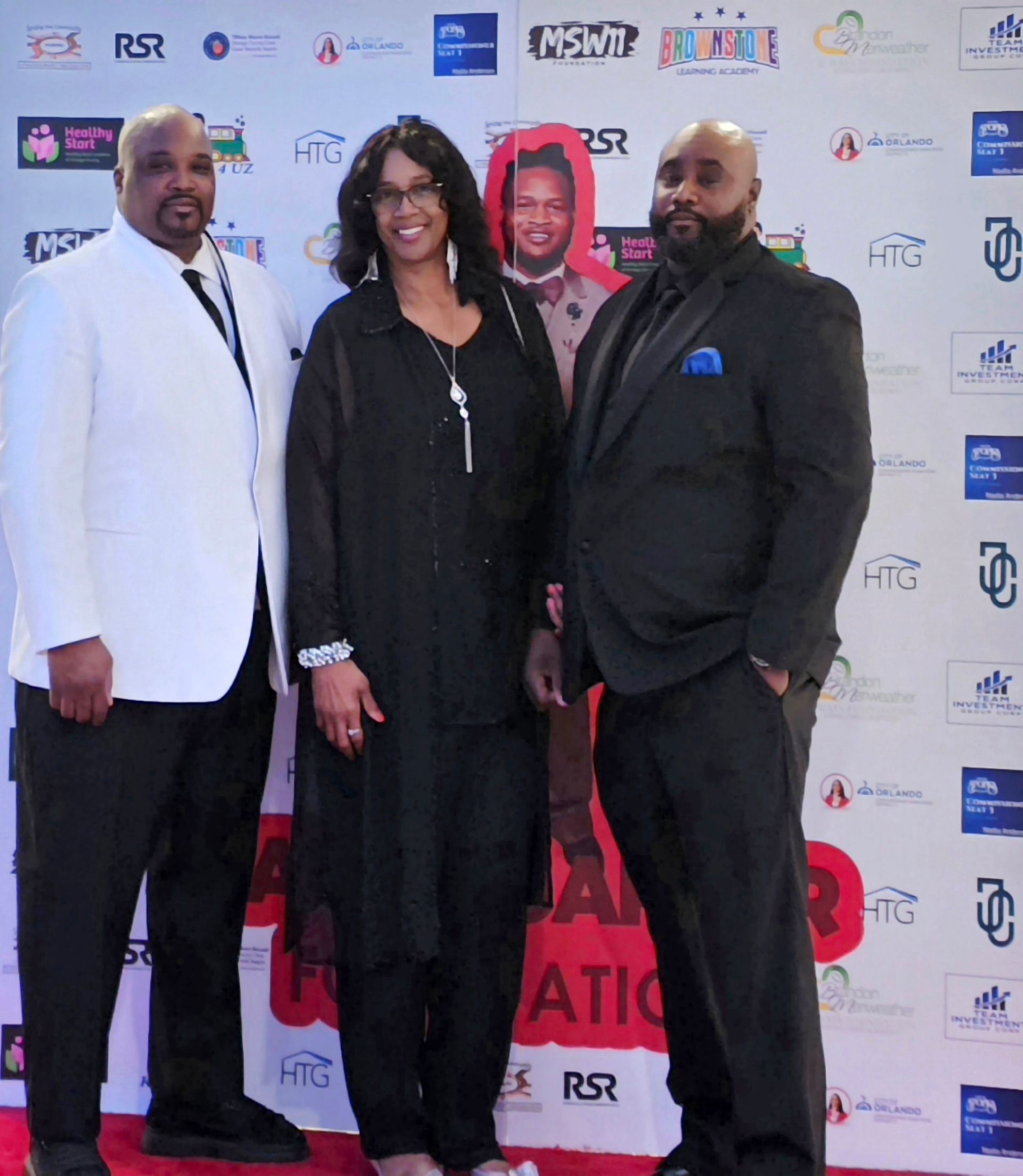 Three people posing on a red carpet in formal black and white attire before a logo backdrop