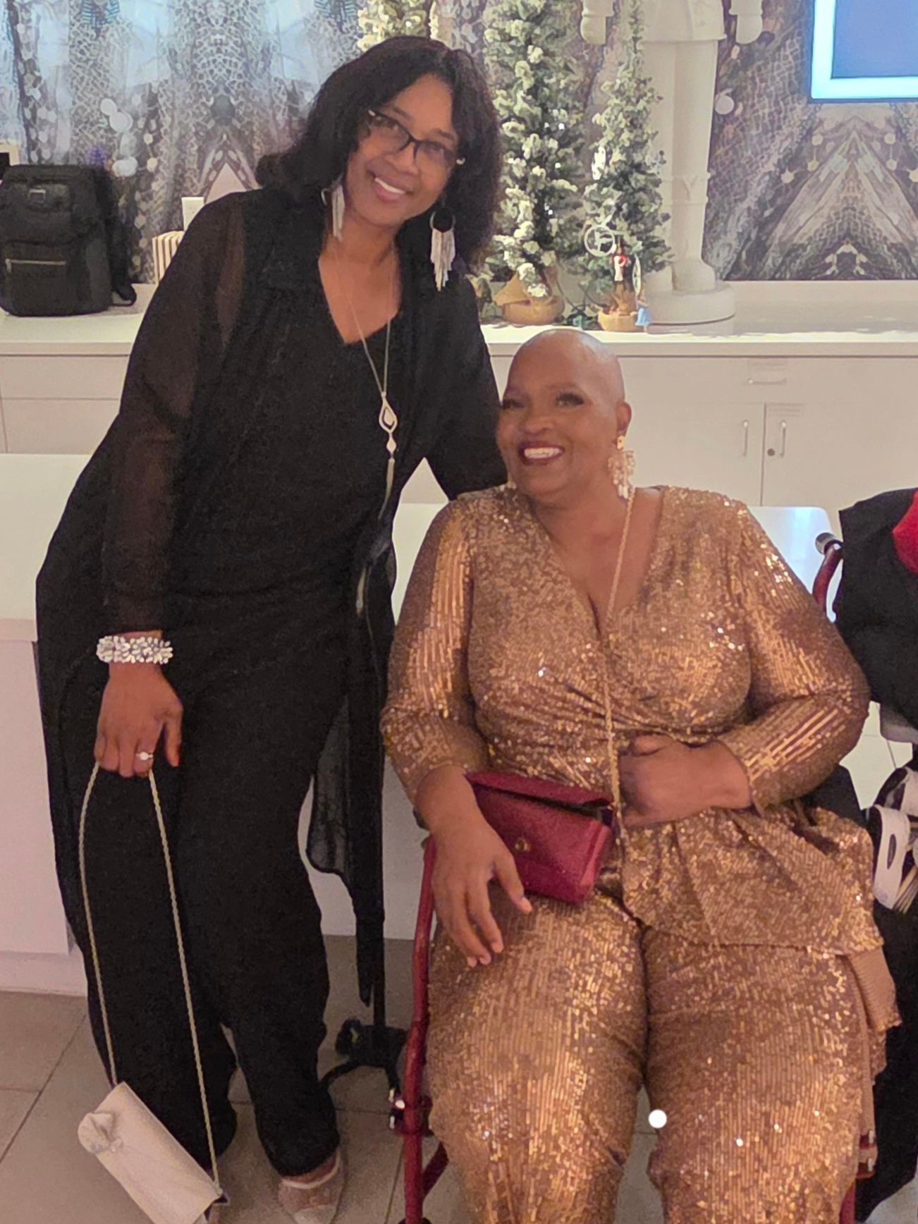 Two women smiling indoors, one standing in black and one seated in a gold dress holding a burgundy clutch.