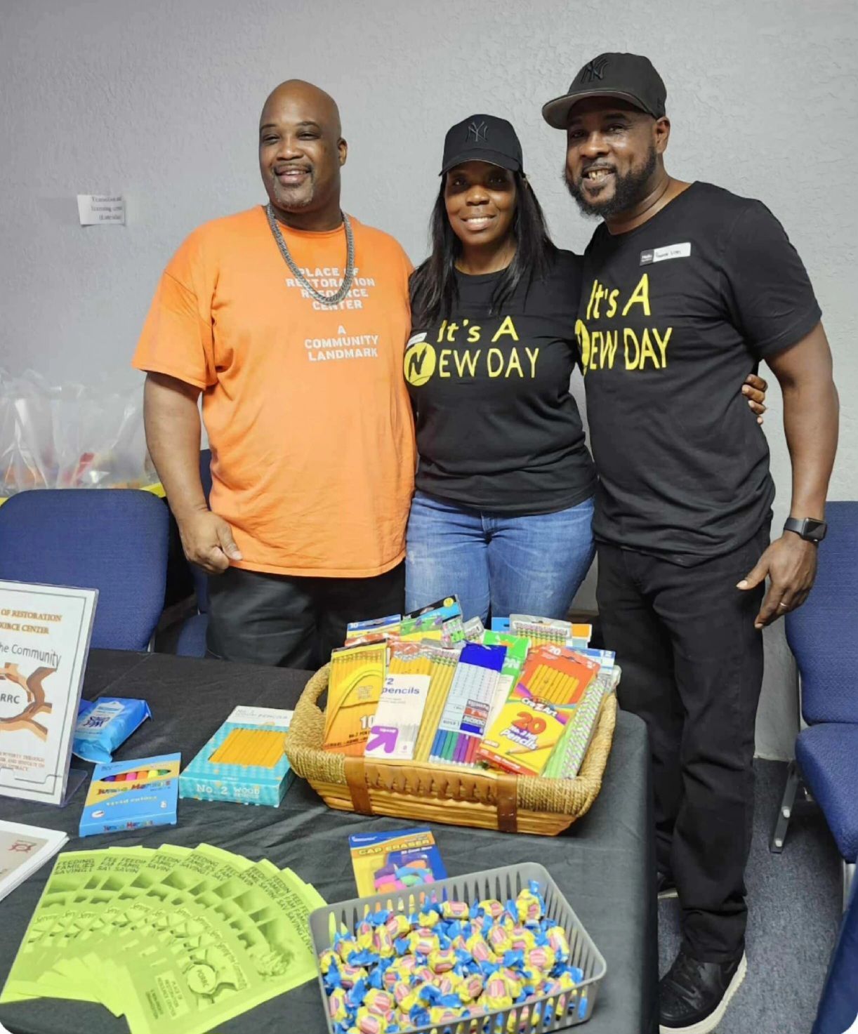 Three people stand behind a table with items. One wears orange, two wear black shirts that say “It’s A New Day.”