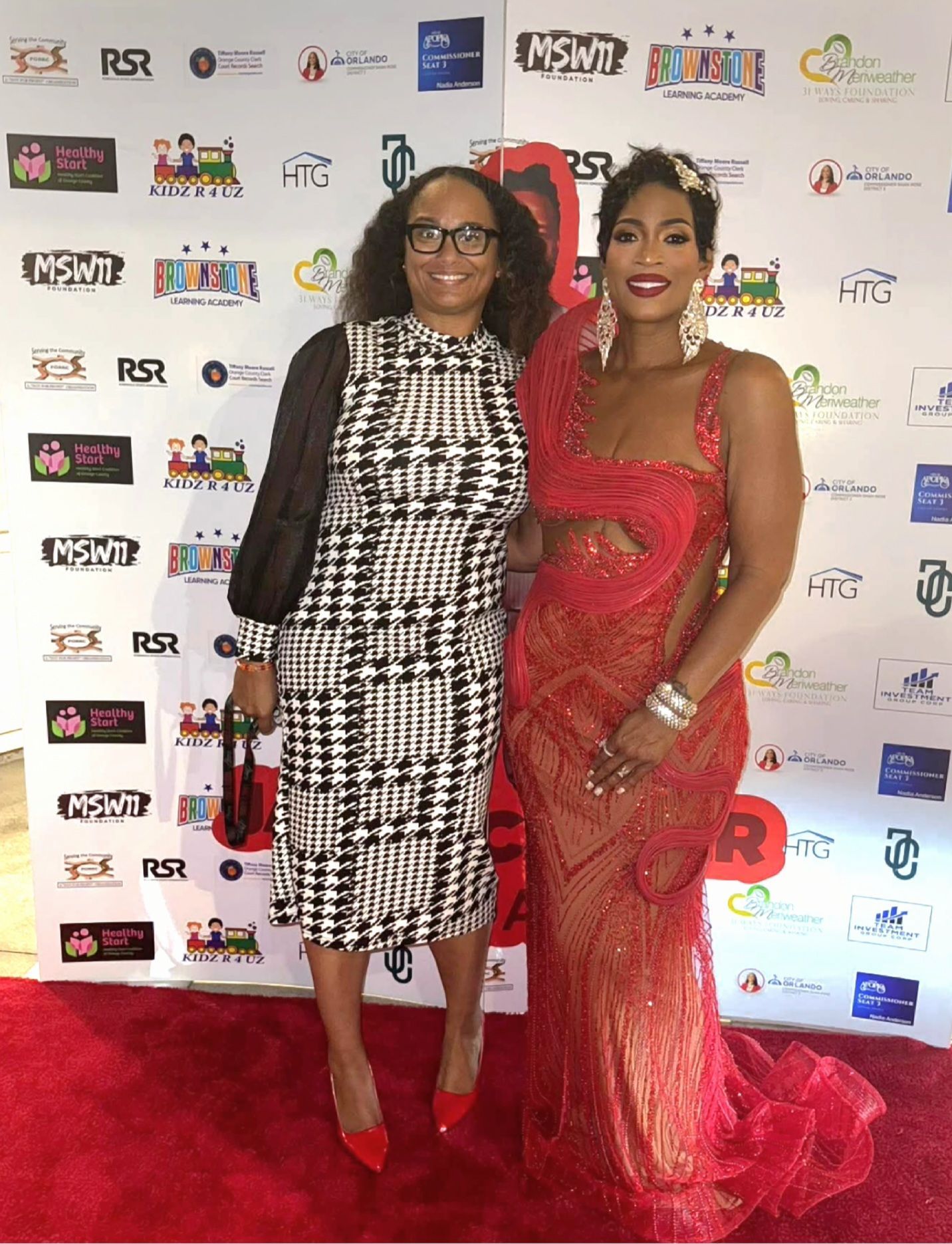 Two women posing at a red-carpet event, one in a black-and-white dress and one in a red gown.