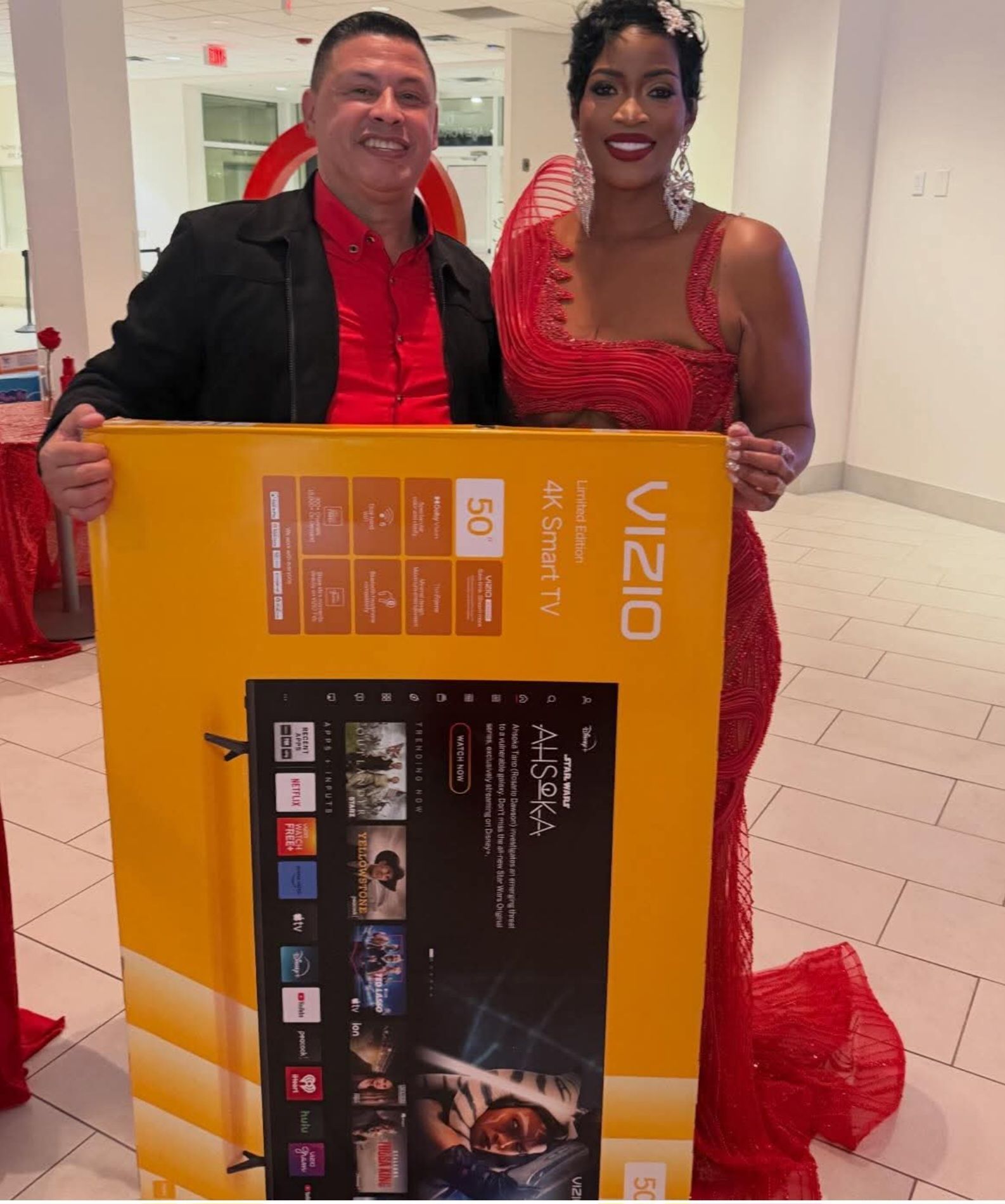 Two people in red attire holding a large Vizio TV box in a bright lobby.
