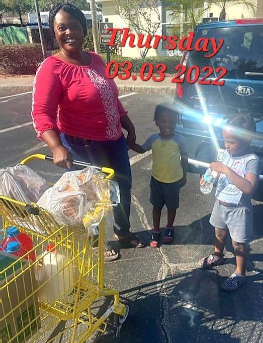 Woman with two children and shopping cart in a parking lot. Cart holds groceries. Sunshine.