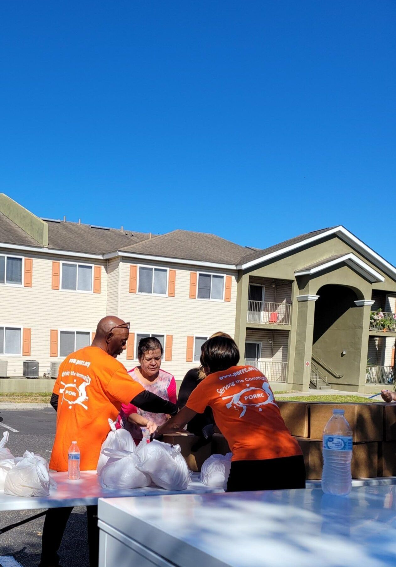 Volunteers in orange shirts distribute food bags at an outdoor table; apartment building in background.