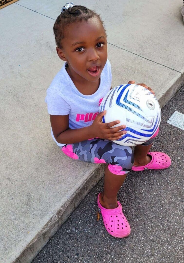 Child holding a soccer ball, sitting on curb, wearing pink Crocs and camo leggings.