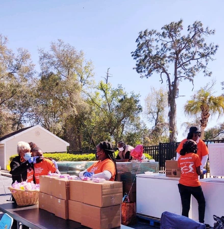 People in orange shirts at outdoor food distribution, near tables and boxes, under trees.