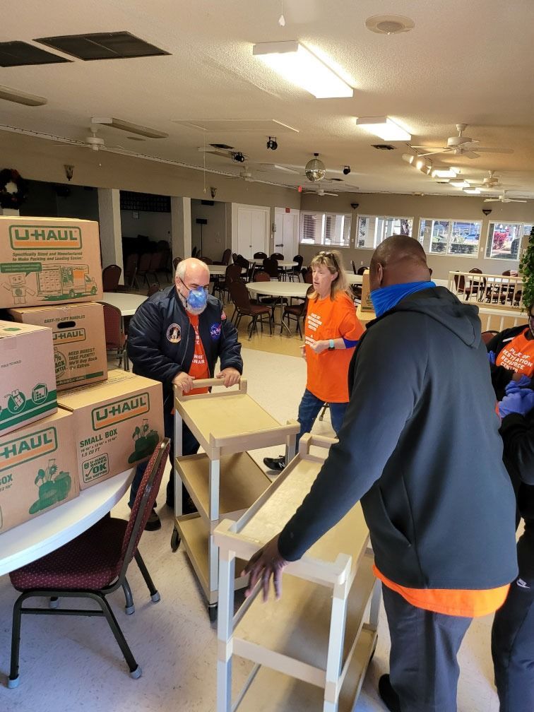 People in a room, moving boxes and shelving. One is wearing a mask, two wearing orange shirts. U-Haul boxes visible.