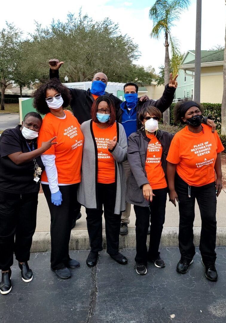 Group of people in orange shirts and masks pose outside. One person raises their fist, another gives a peace sign.