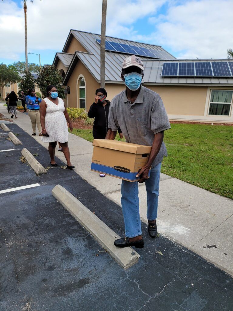 Man in mask carries a box, walking on asphalt. Others queue nearby. Building with solar panels.