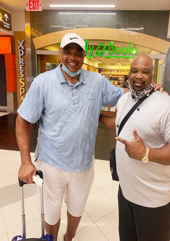 Two men pose in front of a store entrance. One points, the other smiles with a suitcase. Brightly lit mall setting.