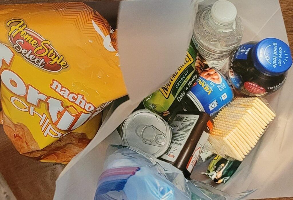 Bag with snacks: tortilla chips, water bottle, juice, crackers, soda, and small plastic containers.