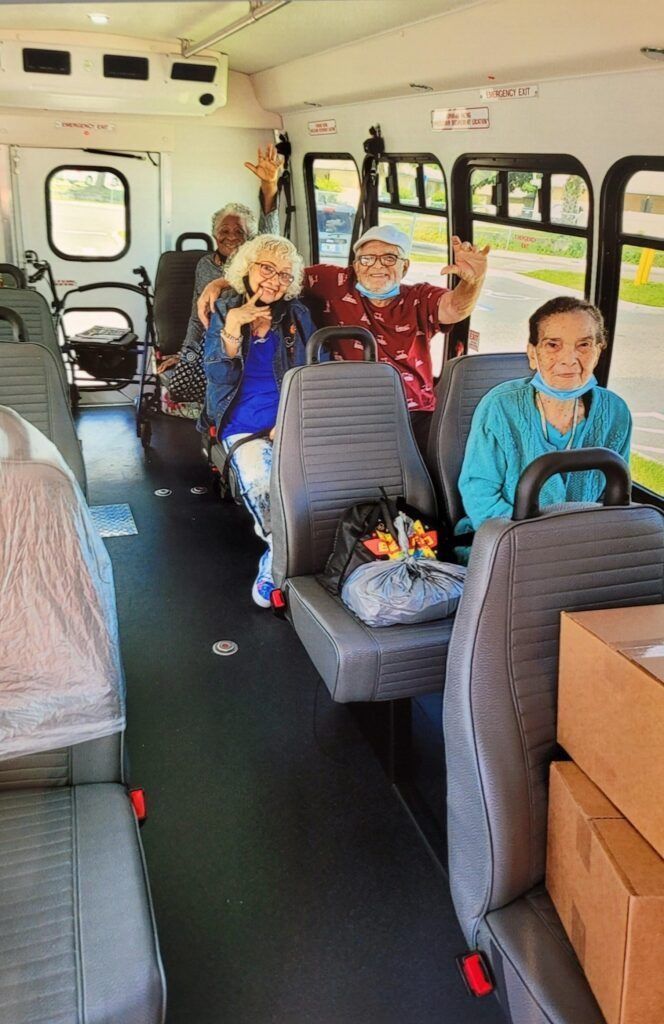 People smiling and waving from the seats of a bus. Boxes and bags are visible.