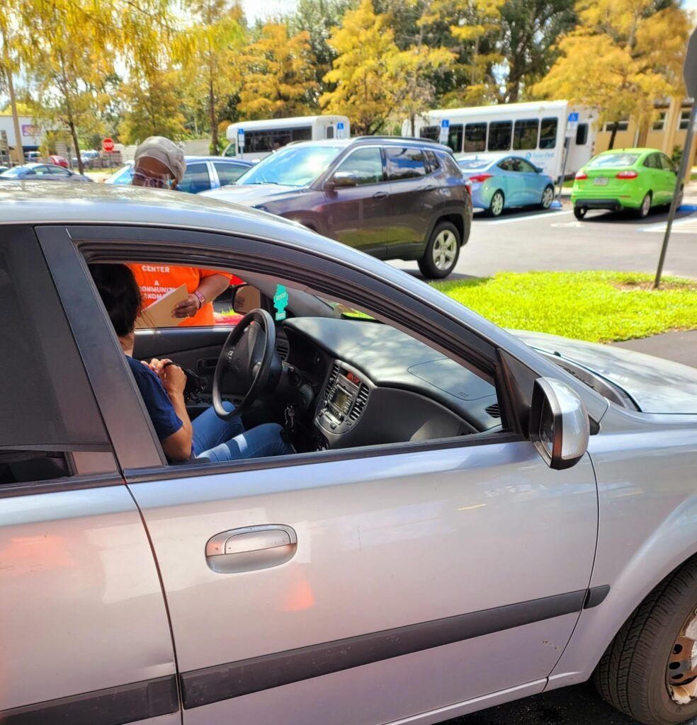 Silver car at a drive-through window. A person is leaning out of the car. Buses and other cars are in the background.