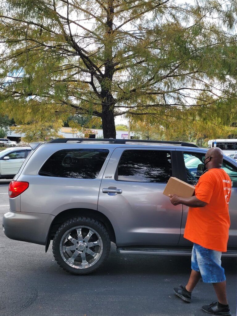 Man in orange shirt with cardboard box next to silver SUV in parking lot. Tree in background.