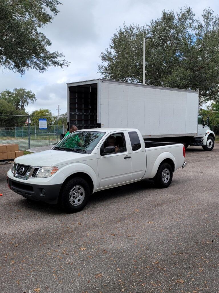 White pickup truck parked in front of a white moving truck; outdoors, overcast sky.