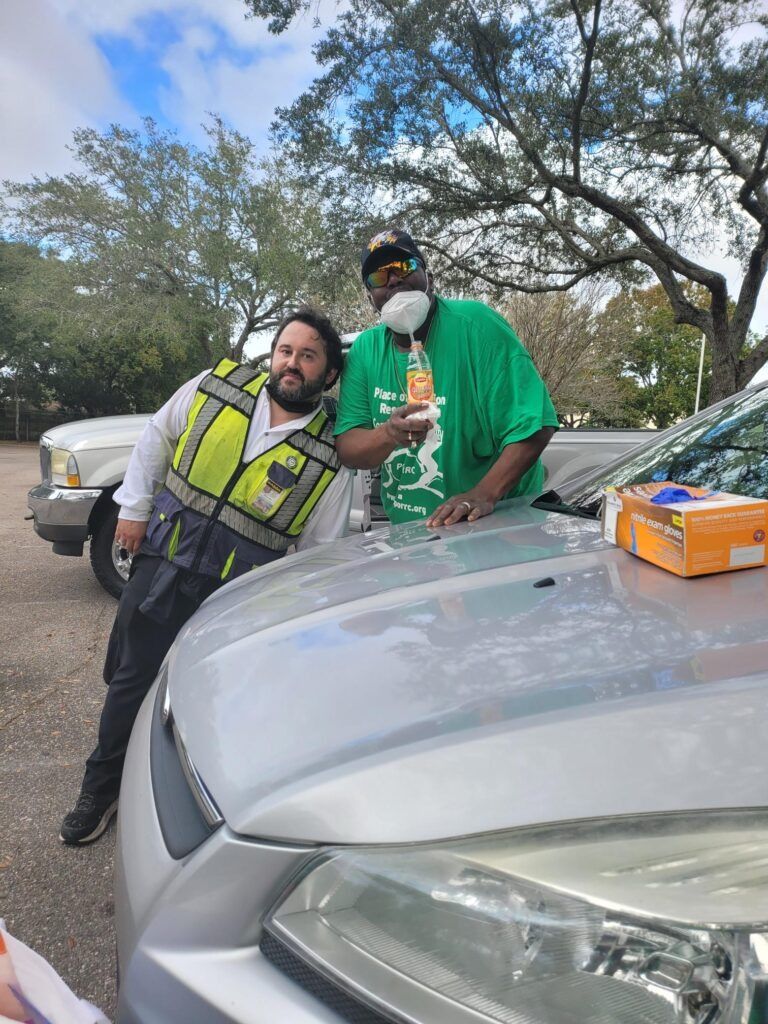 Two people next to a car. One wears a green shirt and mask, the other a reflective vest. Outdoors.