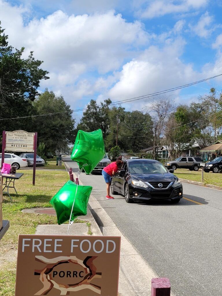 Free food drive: Person hands food to driver in black car. Green balloons mark the location, street scene.