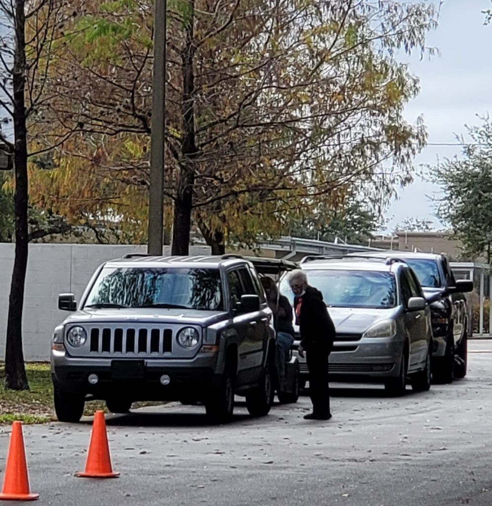 Cars lined up near a building, with people interacting near the vehicles. Orange cones sit in front.