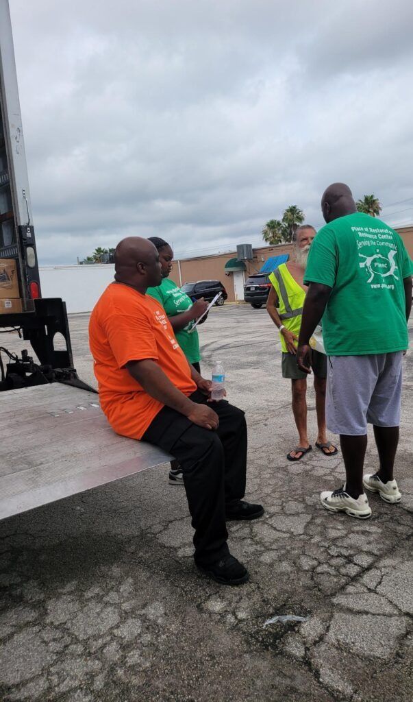 Group of people near a truck, some wearing orange and green shirts. Cloudy sky.