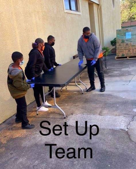 Four people, masked, setting up a black table outside a building.