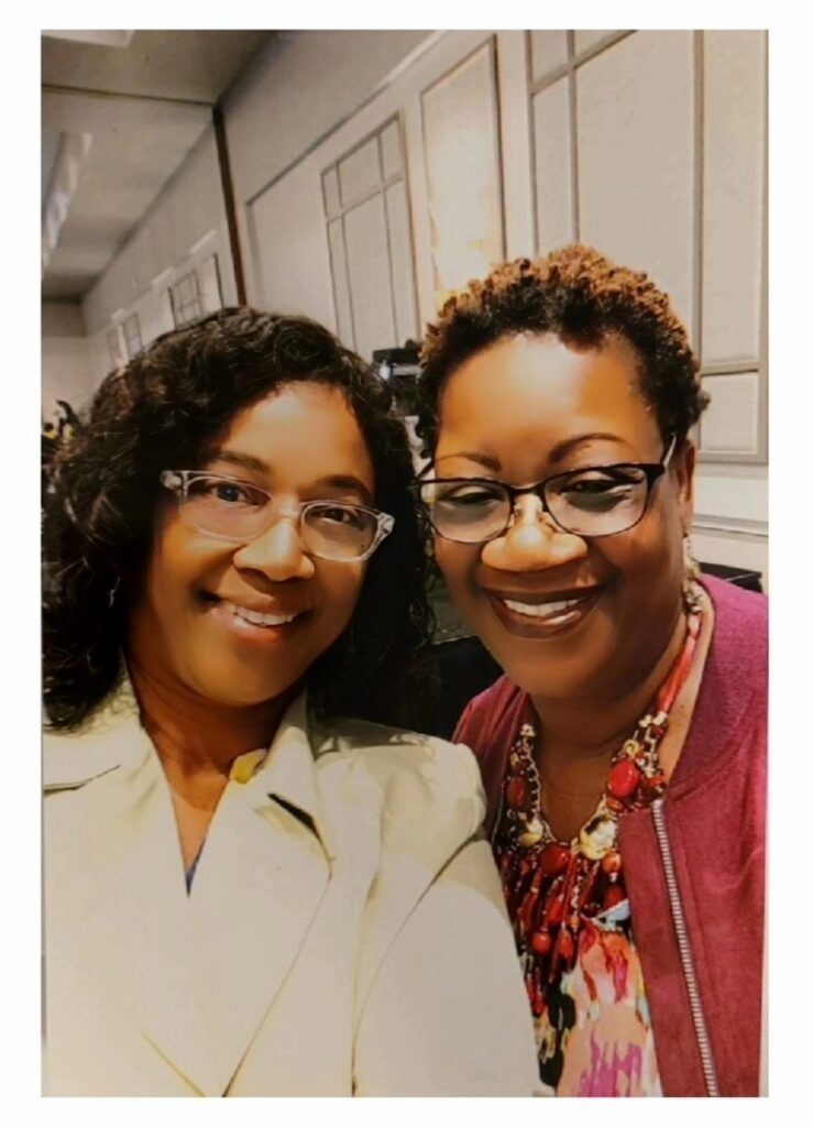 Two smiling women wearing glasses pose for a selfie indoors. One in a light blazer, the other in a burgundy jacket.