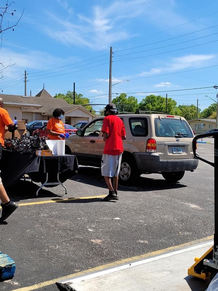 People at a drive-thru food distribution site. Person in red shirt receives items from a table next to a car.