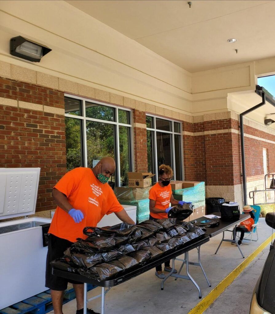 Two people in orange shirts and masks preparing food bags outside a brick building.