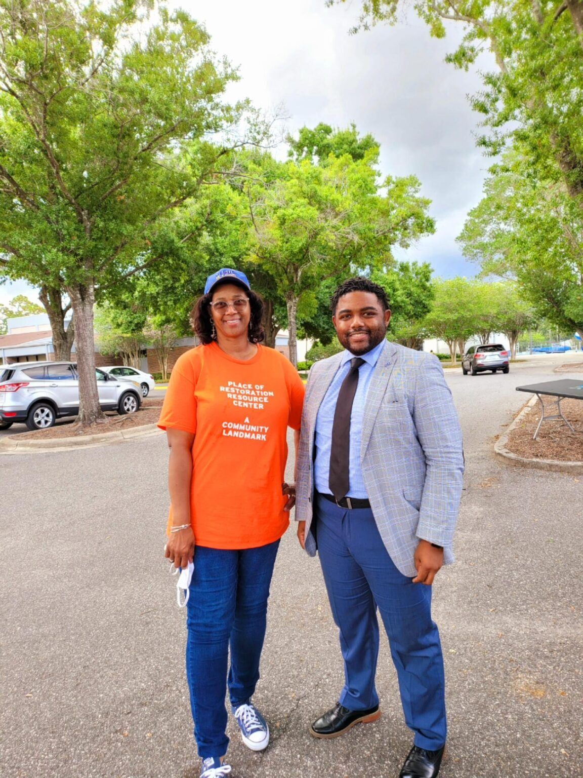 Two people standing outside under trees, one wearing orange shirt, the other a blazer.