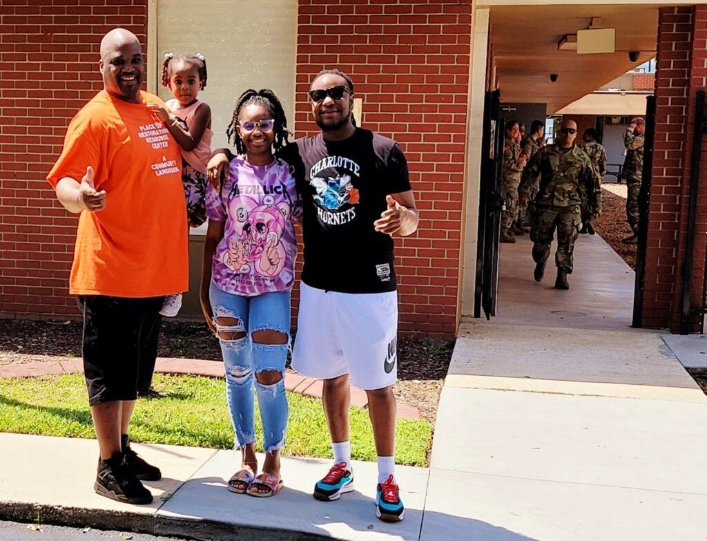 Family poses outside a brick building as soldiers walk by. Man in orange holds a child, two others smile.