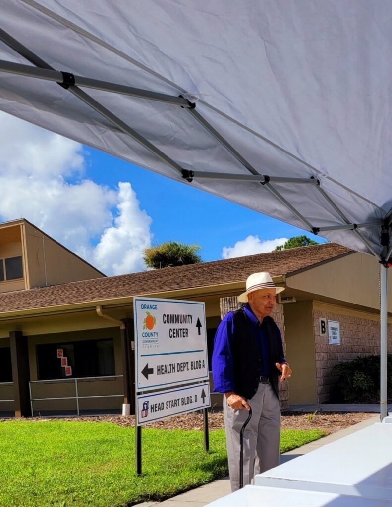 An elderly man with a cane under a tent near a building, likely a polling place.