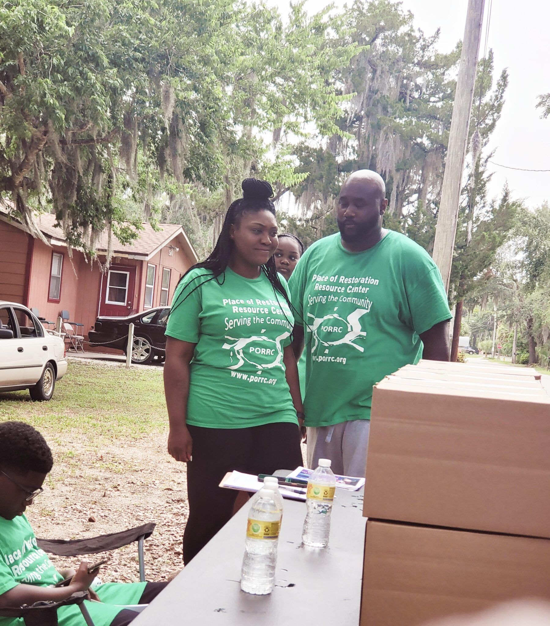 People wearing green shirts stand near a table outdoors; buildings and trees are in the background.