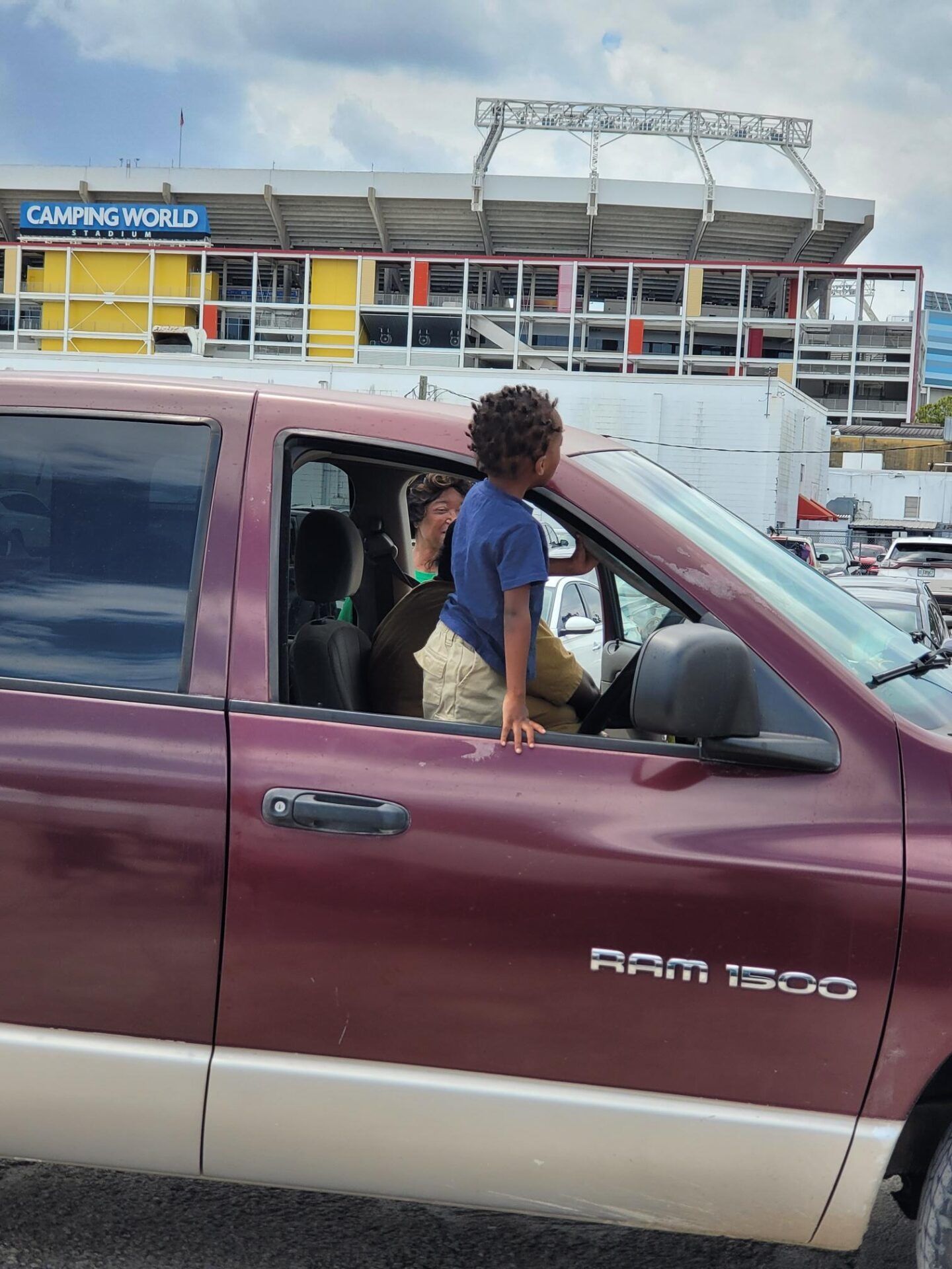 Person and child in a maroon Ram 1500 truck with Camping World Stadium in background. Child leaning out window.