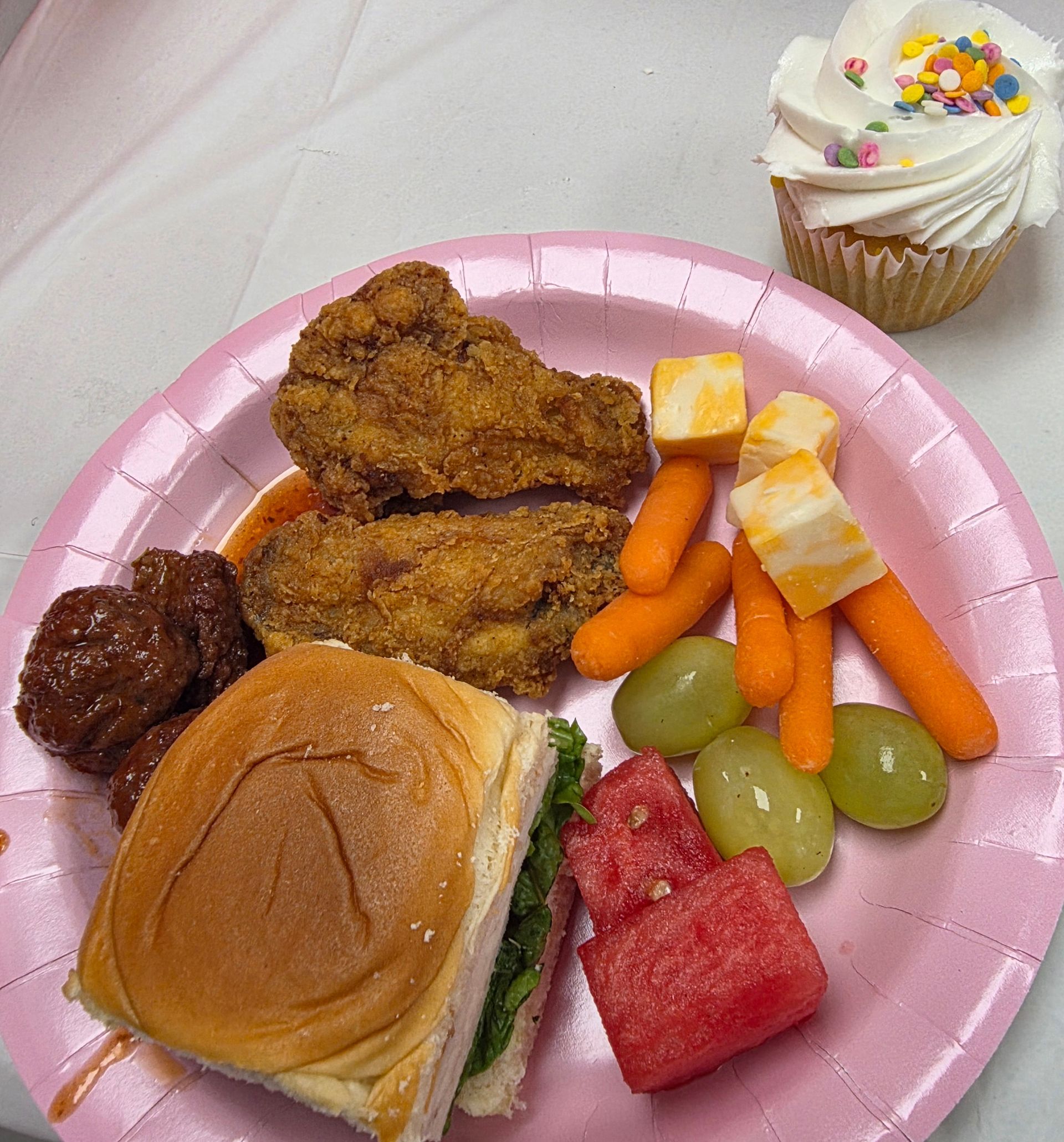 Pink plate with fried chicken, sandwich, carrots, grapes, watermelon, and a cupcake on a white table