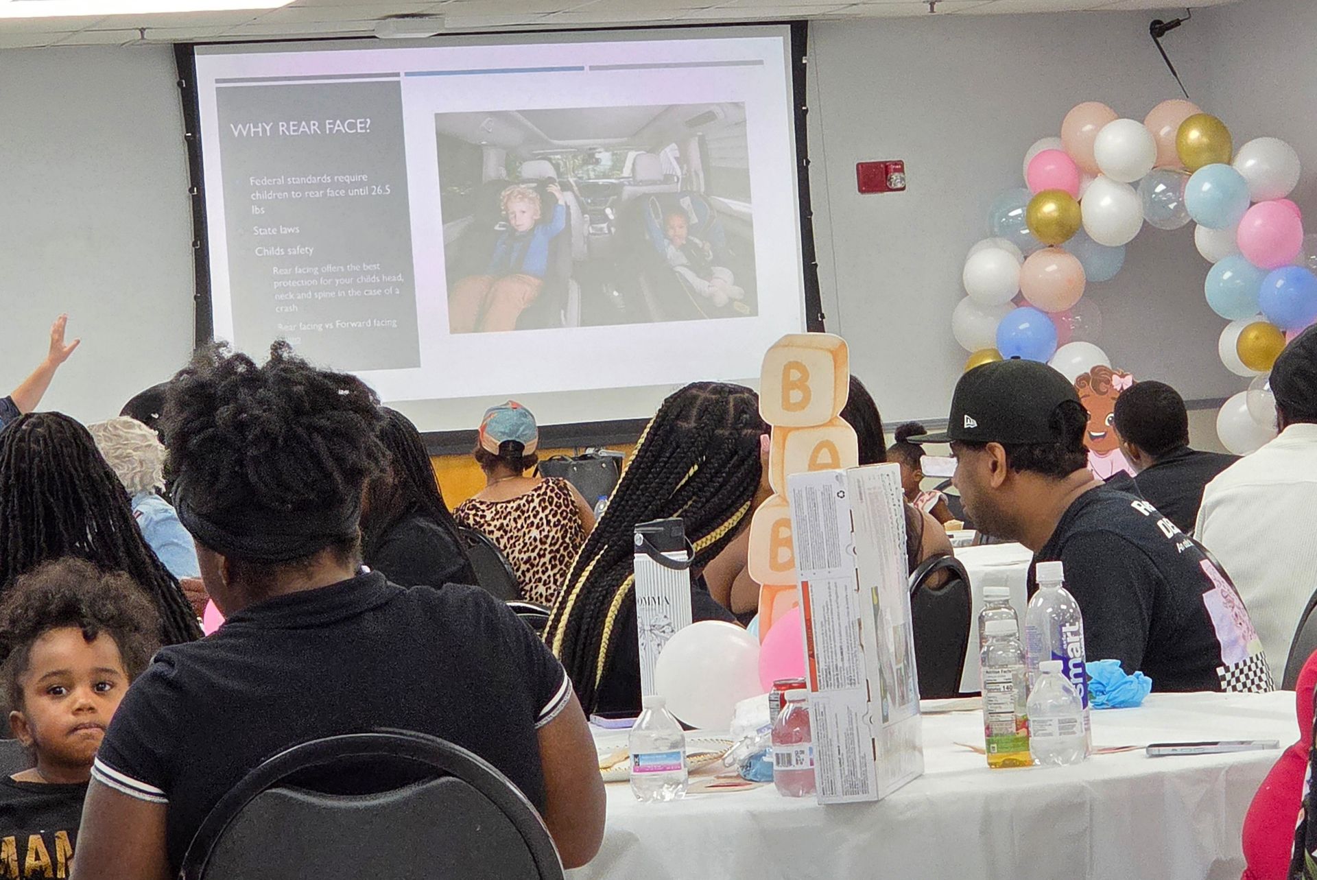 Audience seated at tables in a decorated hall, facing a presentation screen and balloon arch.