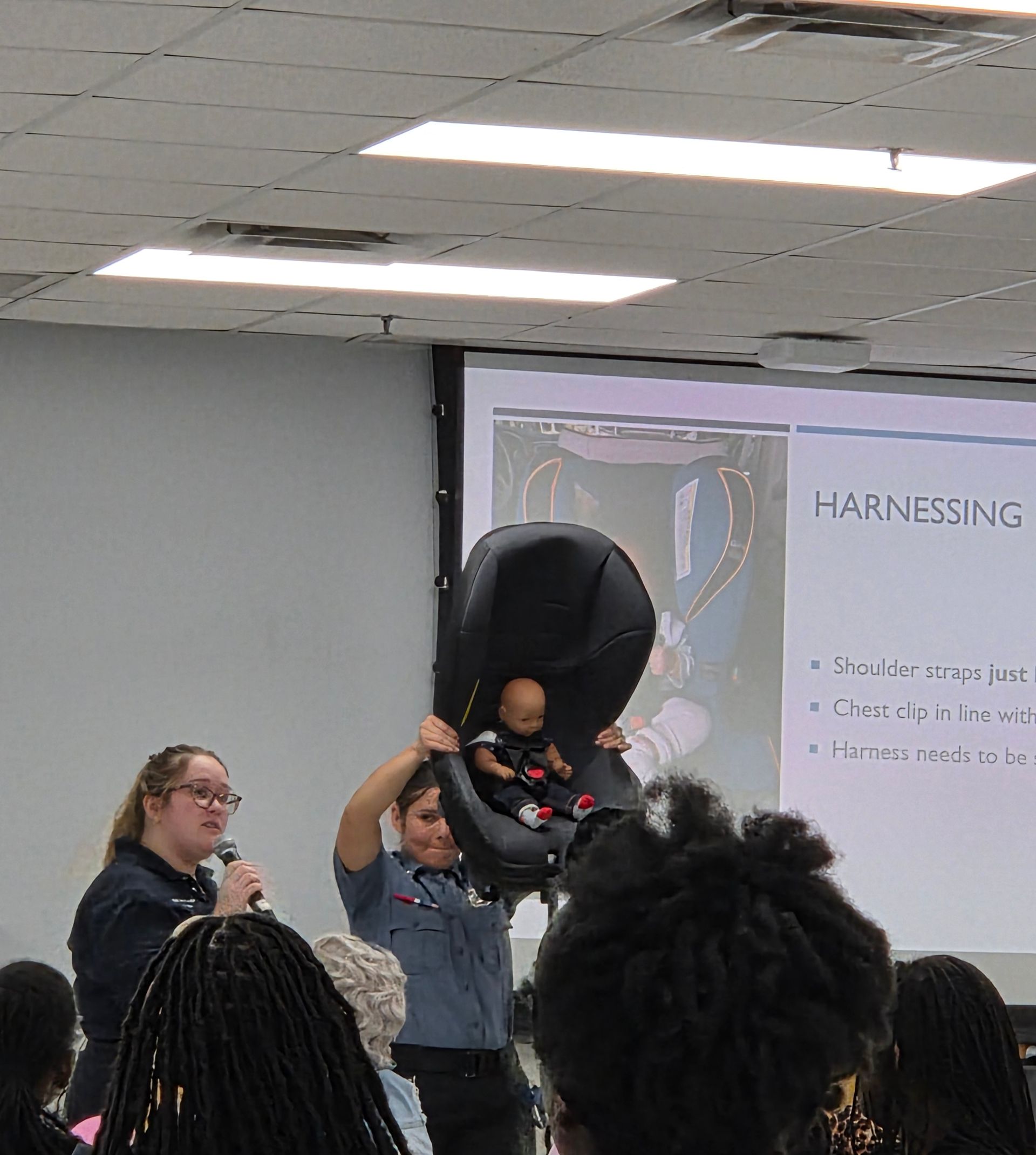 Indoor presentation with a speaker and another person holding up a large black baby seat in front of a projected slide.