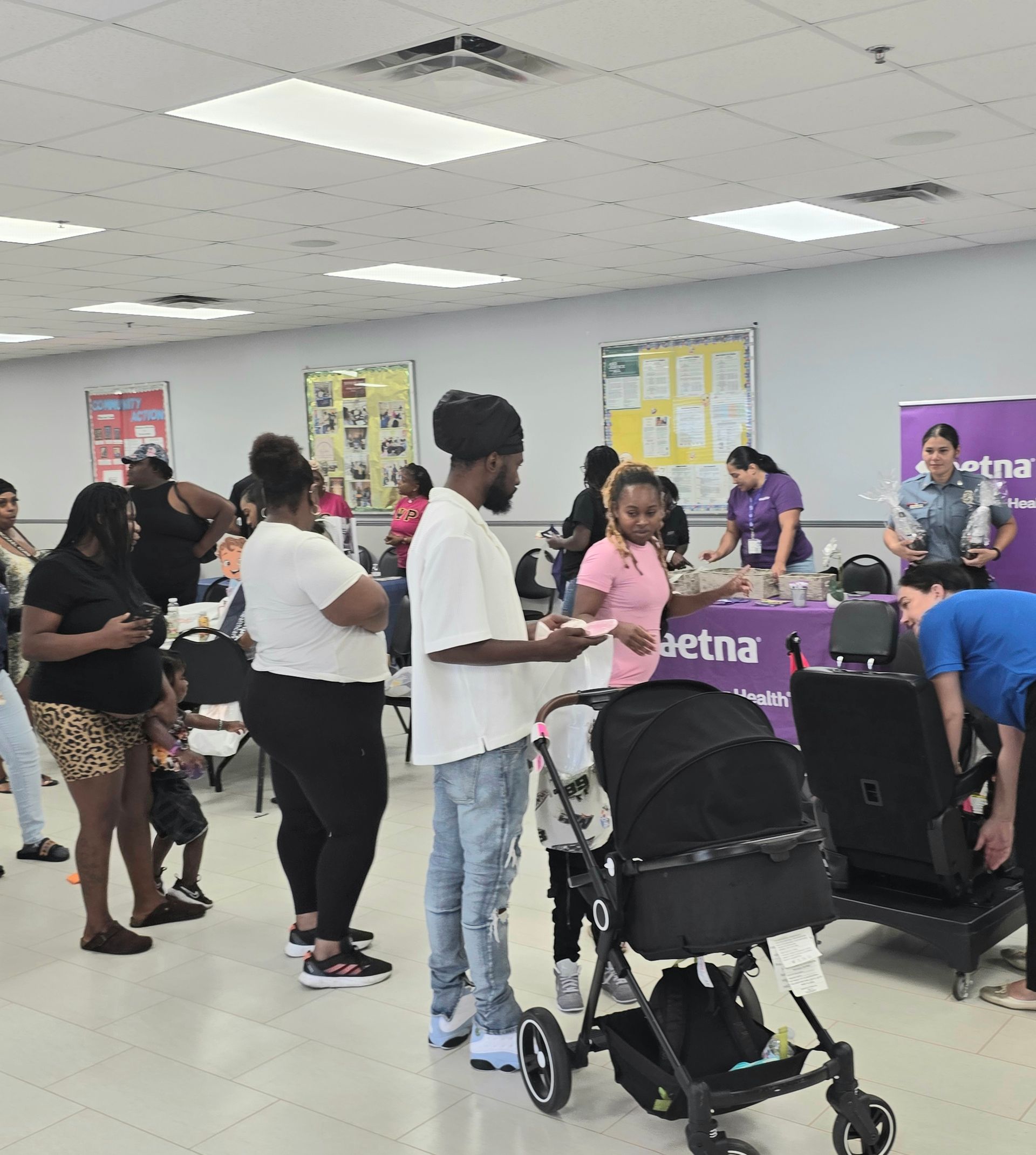 People standing in a community room near purple tables, with a stroller in the foreground.