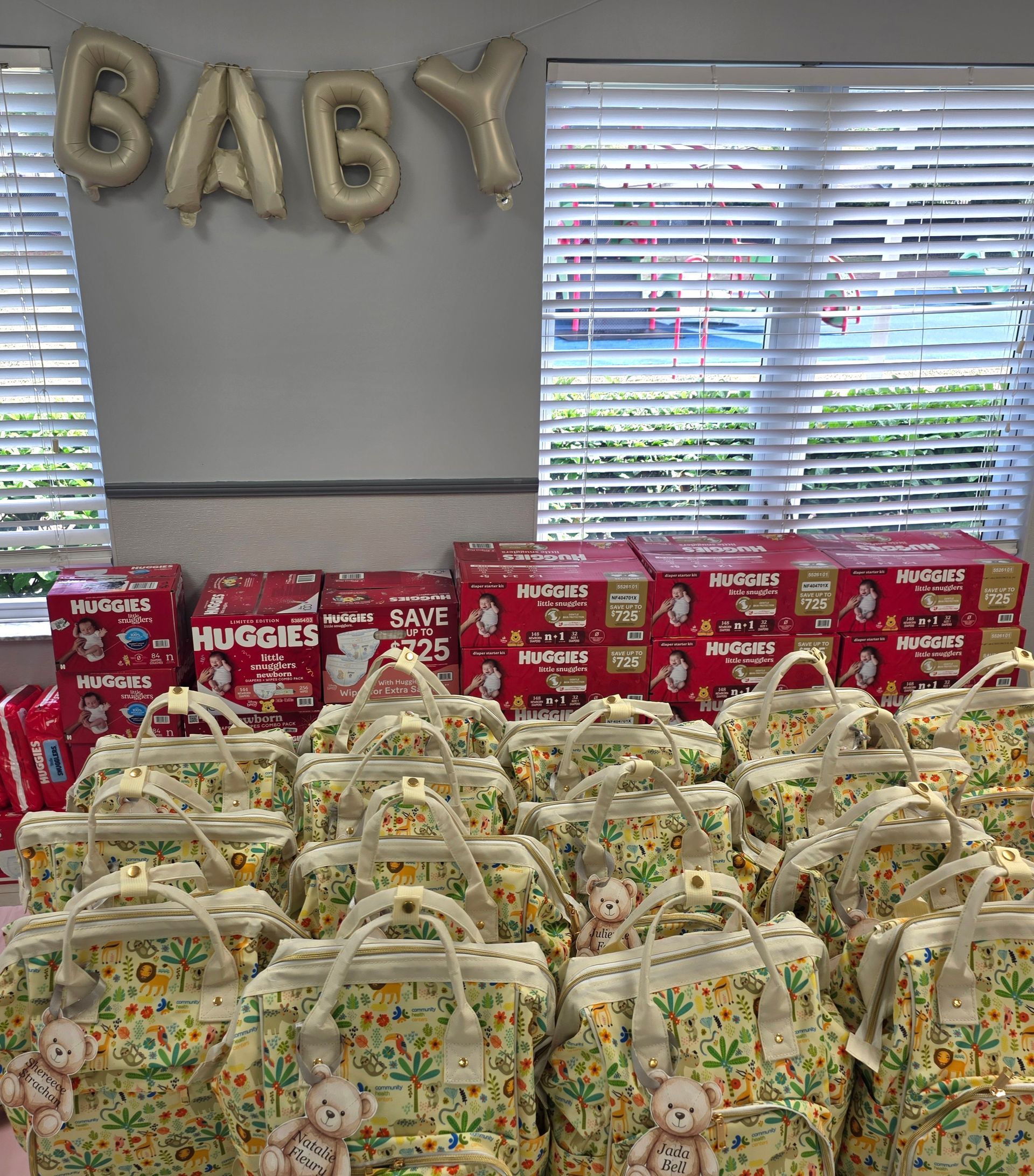 Baby shower gift display with gold balloons, stacks of diapers, and patterned baby bags in front of windows
