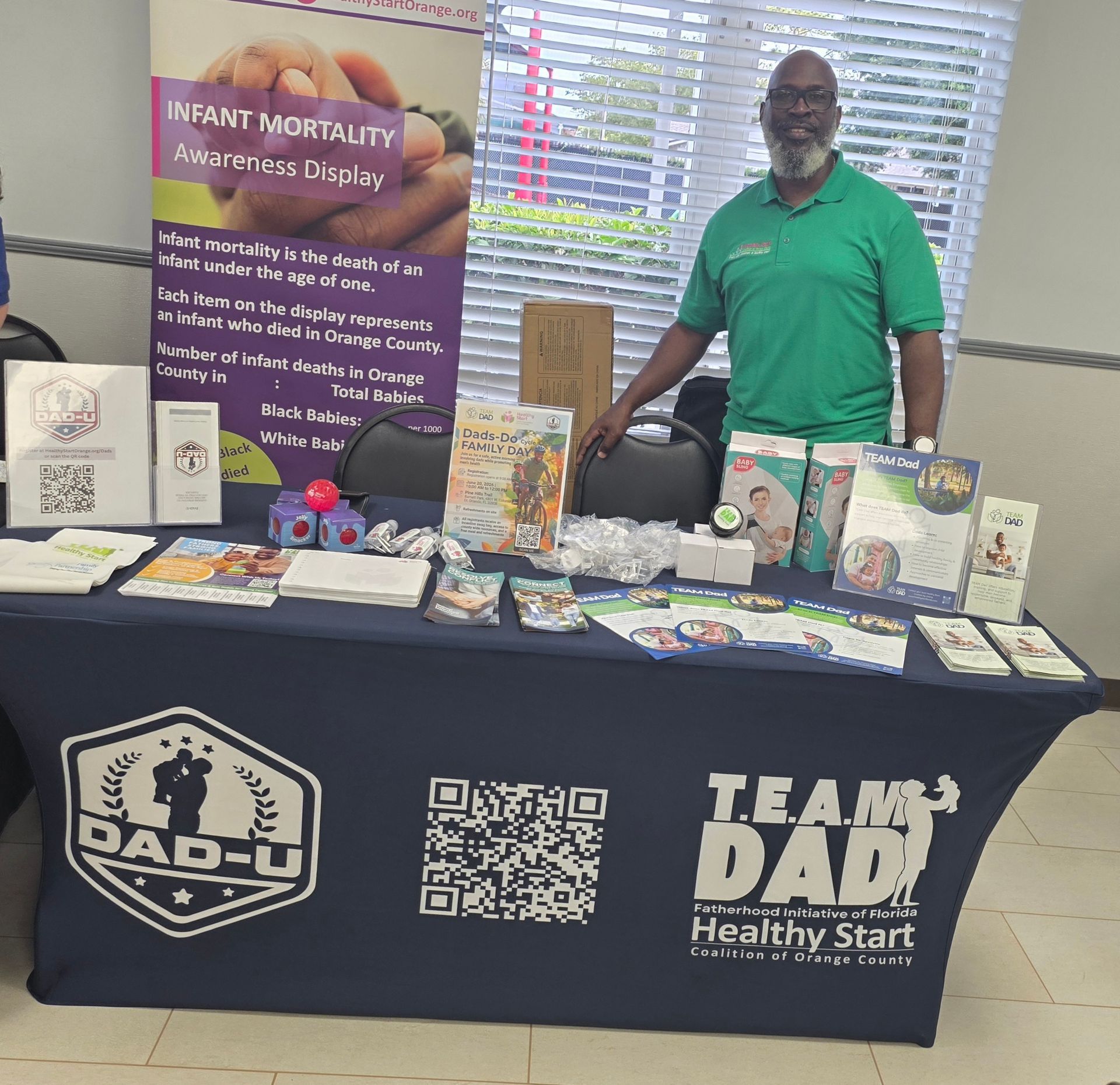 Man standing behind a Team DAD display table with brochures, gifts, and a purple banner at an event.