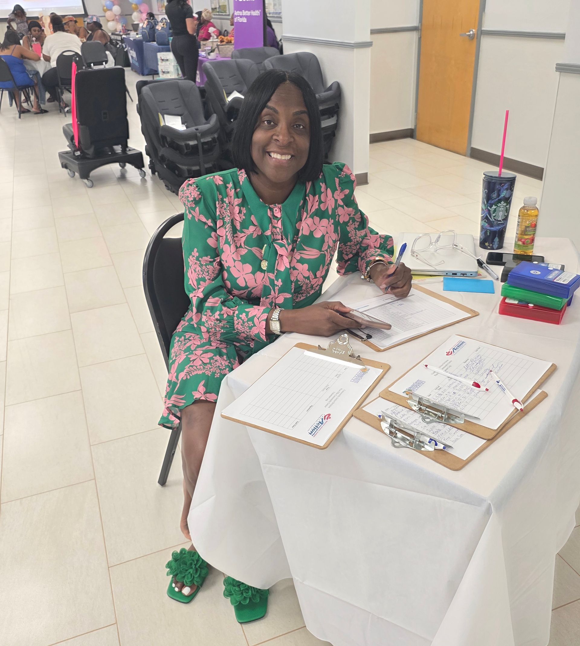 Woman at a table smiling while signing papers at an indoor event booth, with flyers and a drink nearby.