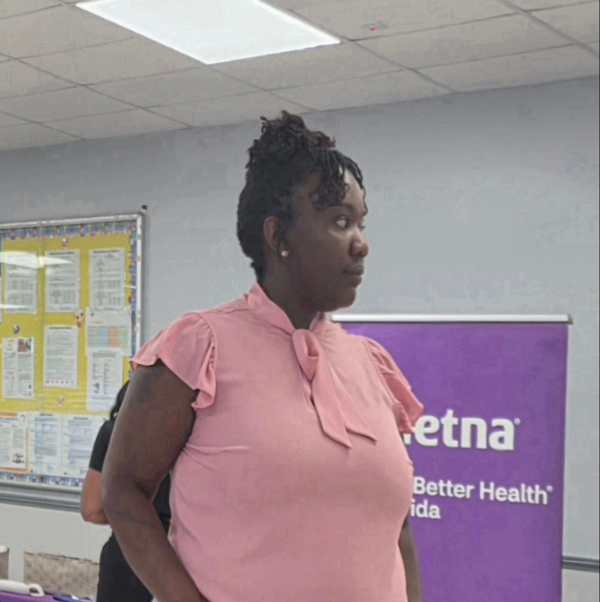 Woman in a pink blouse standing indoors beside an Aetna banner and bulletin board.