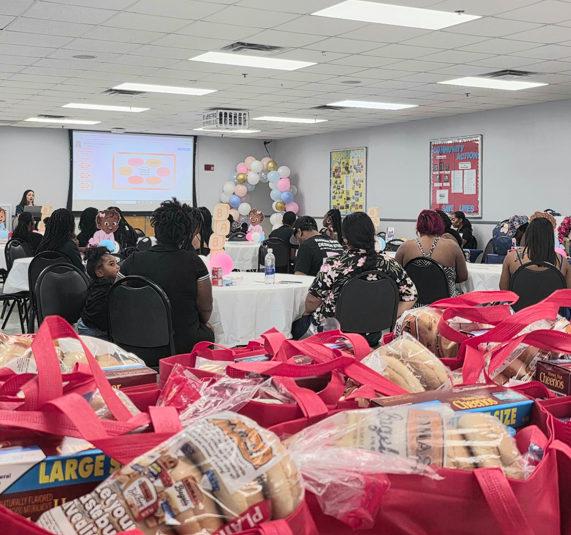 Community event in a decorated hall, with seated attendees and pink gift bags in the foreground.